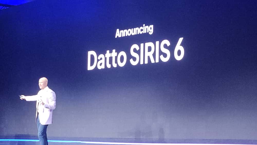 A speaker presents on stage at DattoCon 2025 with a large screen showcasing the launch of Datto SIRIS 6, highlighting features such as flexibility to expand, small form factor options, and industry-best RPO value.